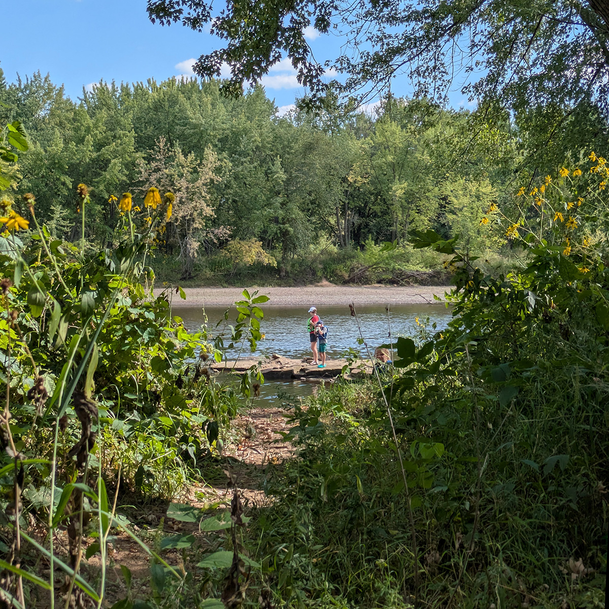 A cozy path leads down to the river. Bean Island is in the background. Two people are in the distance at the waterfront.